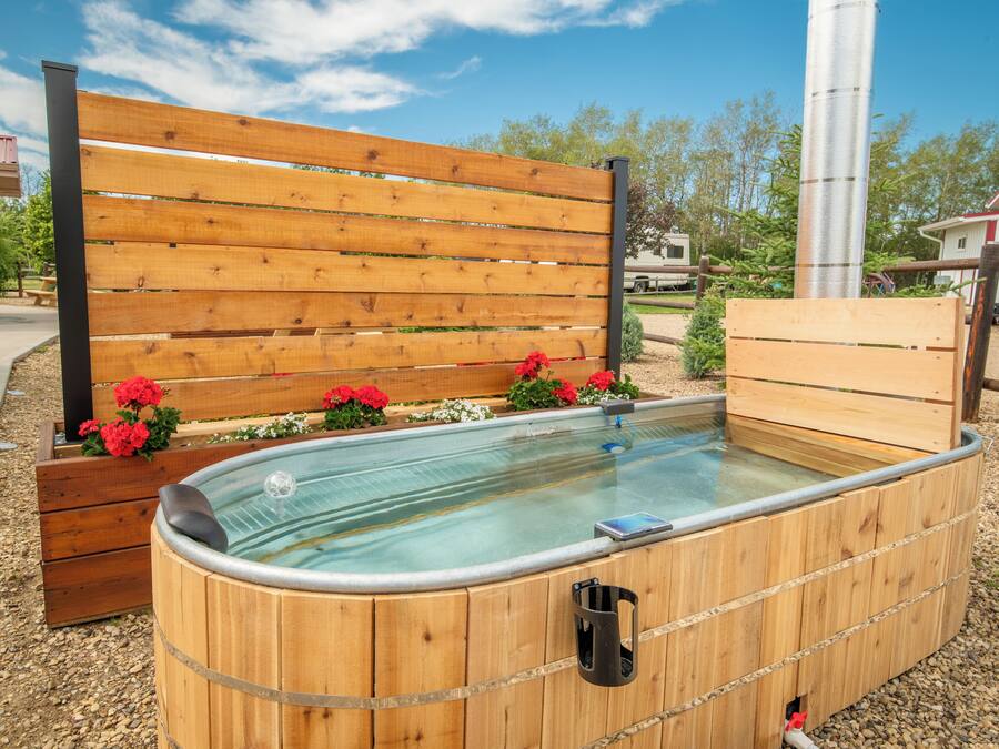 A wooden hot tub with a bench and flowers, surrounded by gravel and trees.