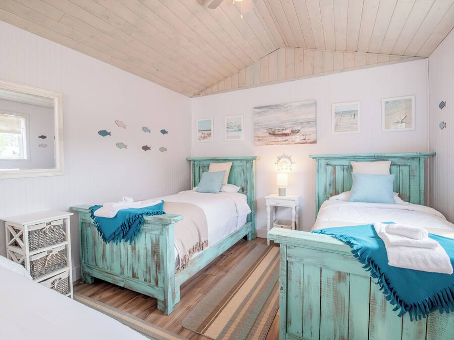 Una habitación con dos camas, pisos de madera, un espejo, decoración en la pared y una pequeña mesa con lámpara.