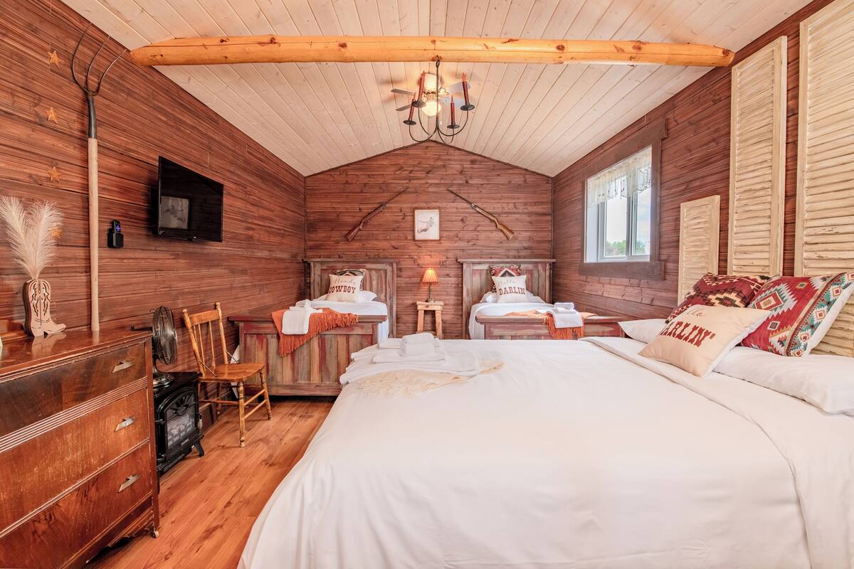 A cozy room with a large bed, two bedside tables, a wooden dresser, a vintage stove, and a television on the wall.