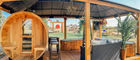 A wooden deck with a sauna, outdoor seating, and a view of a red circular structure in the background.