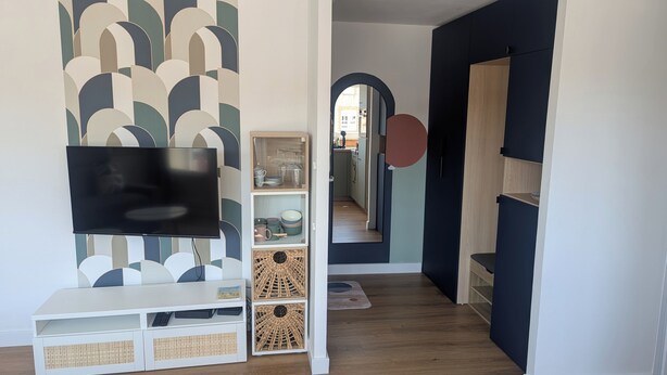 Un salon moderne avec un téléviseur à écran plat fixé au mur, un meuble de rangement blanc agrémenté d’un panier en osier, et une cuisine visible à travers une arche.