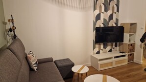 Un salon moderne avec un canapé gris, une petite table ronde, un téléviseur posé sur un meuble blanc et un mur orné d’un motif.