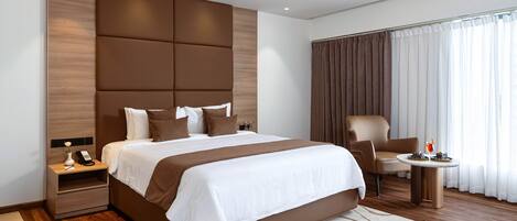 Une chambre d’hôtel moderne avec un grand lit, une table de chevet avec un téléphone, une chaise, une petite table avec un vase et une grande fenêtre avec des rideaux.