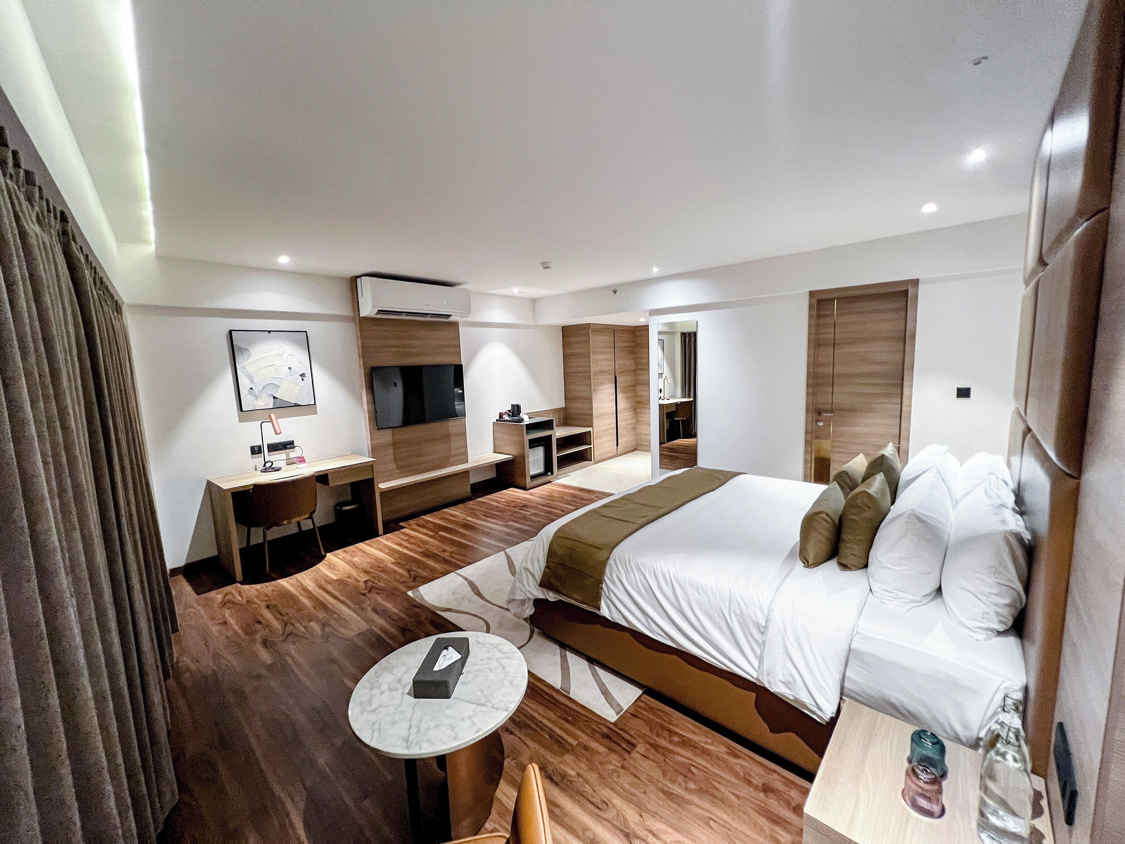 A modern hotel room with a large bed, a desk, a chair, a small table, a TV, and a bathroom visible through an open door.