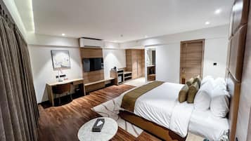 A modern hotel room with a large bed, a desk, a chair, a small table, a TV, and a bathroom visible through an open door.