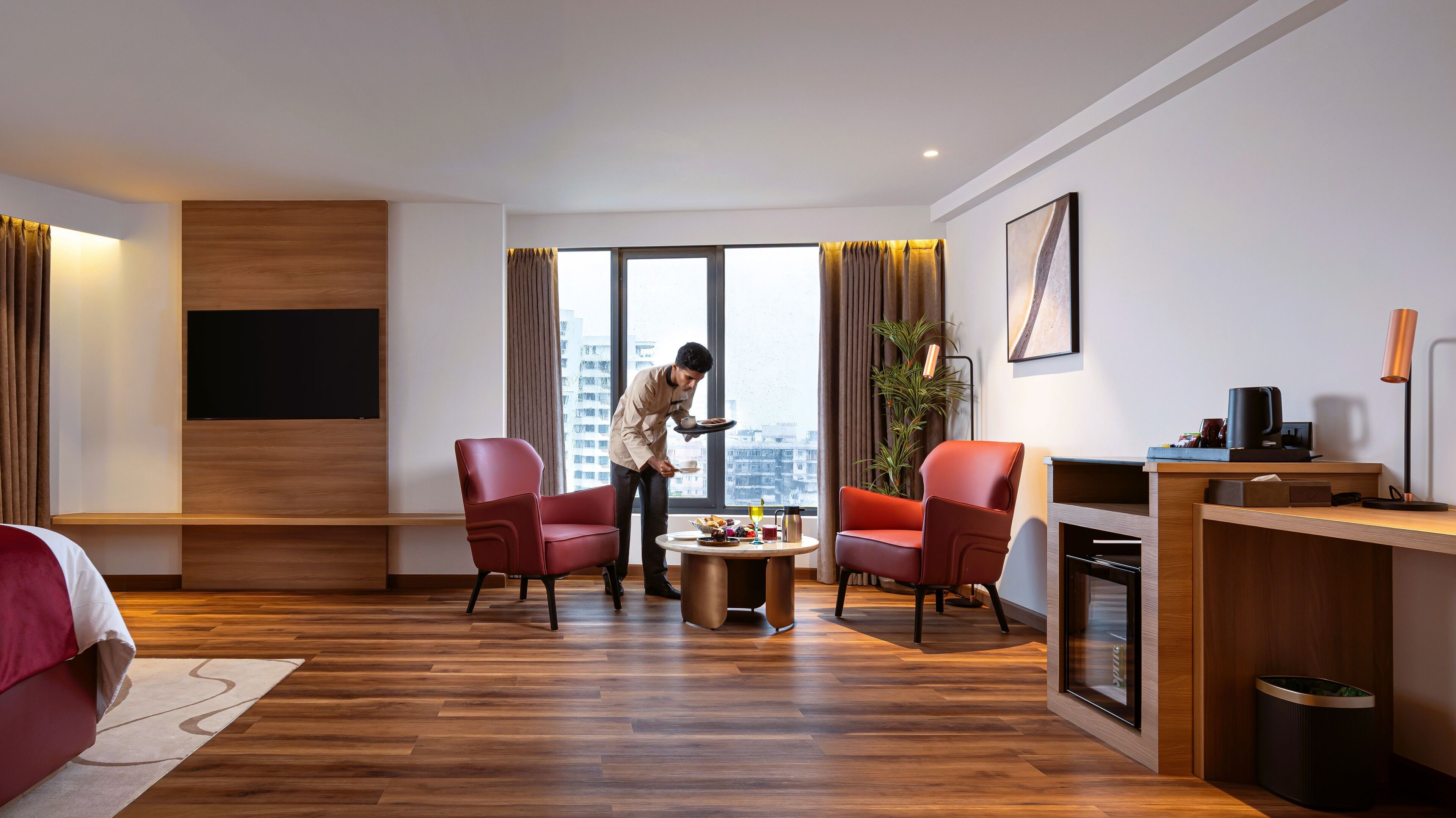 Een moderne hotelkamer met een houten vloer, een eettafel, rode stoelen, een televisie en een groot raam met gordijnen.