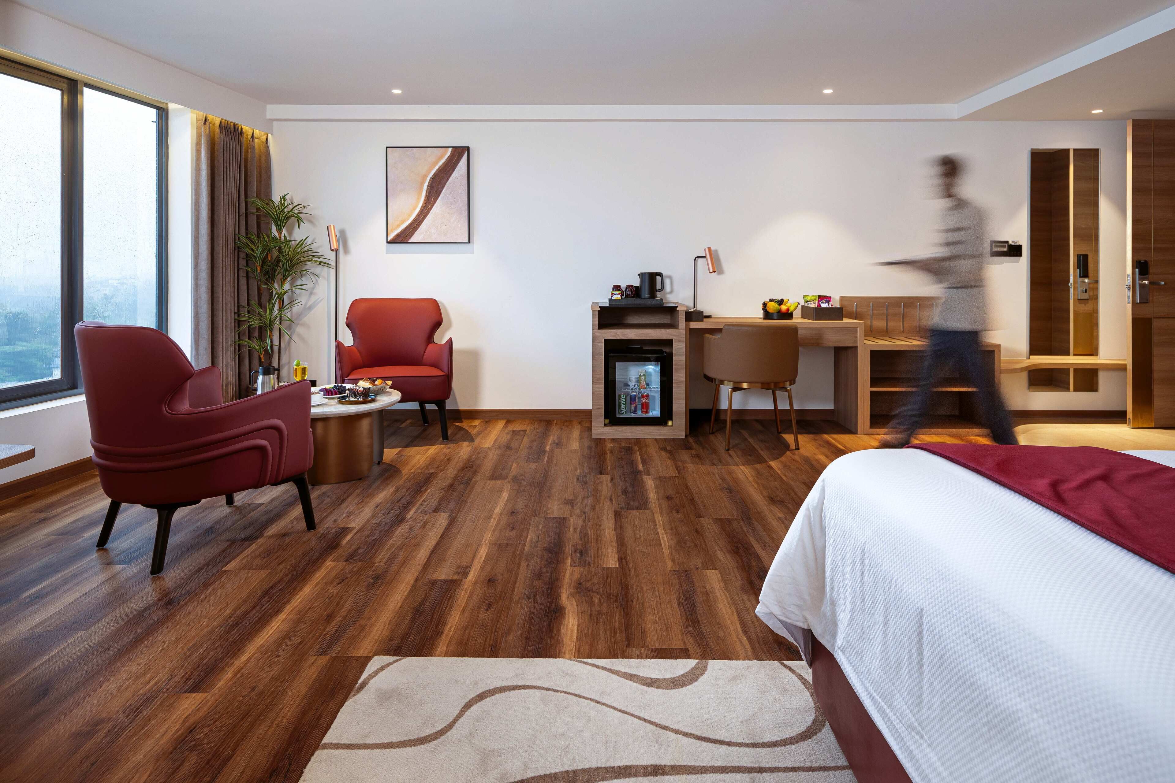 A modern hotel room with a wooden floor, a bed, a desk with a computer, a chair, and a large window.