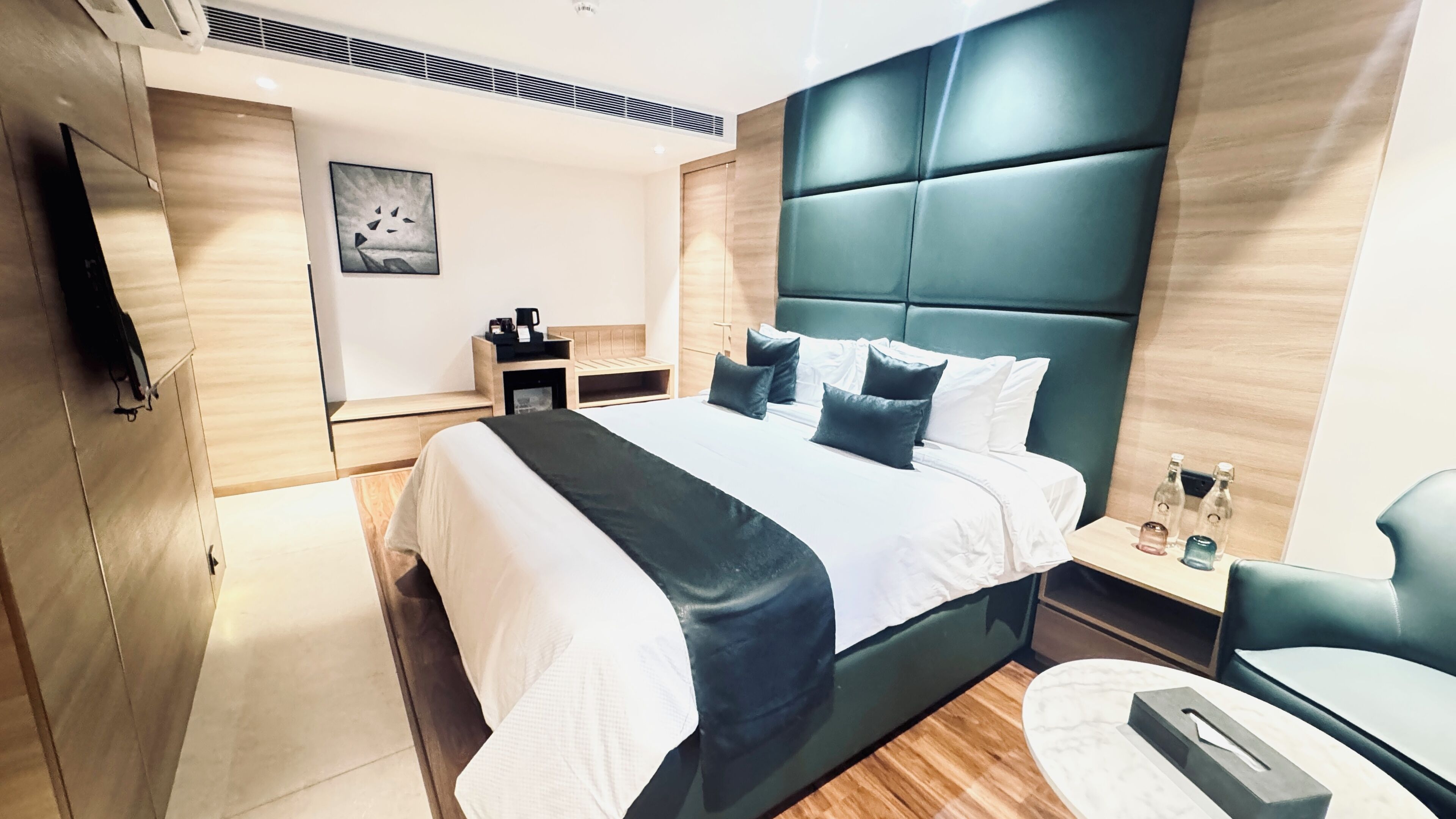 A modern hotel room with a large bed, a bedside table with a lamp, a small seating area, and a wall-mounted TV.
