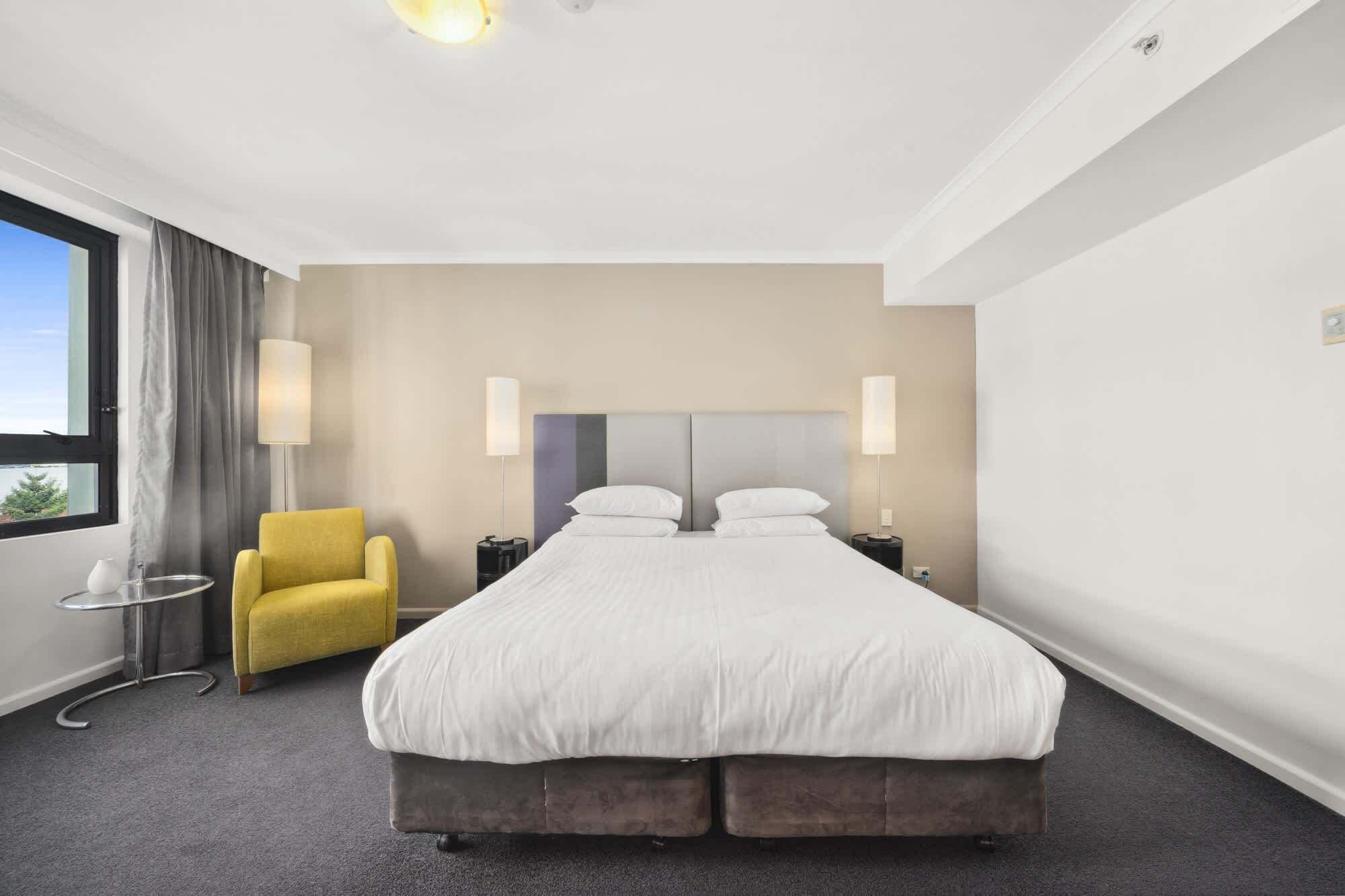 Ein Hotelzimmer mit einem großen Bett, zwei Nachttischlampen, einem gelben Sessel, einem kleinen Tisch und einem Fenster mit Vorhängen.