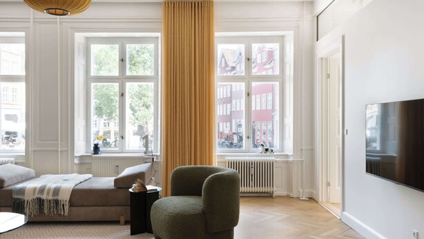 A living room with a sofa, ottoman, and armchair, large windows with a view of buildings, a radiator, and a flat-screen TV. - Former Dukes Residence - 4 bedrooms - 2 bathroms (København)