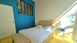 A bedroom with a bed, bench, nightstand, and a window with blinds. - LES LOGIS DU CHATEAU MARIE VICTORIA on the way to ST-JACQUES-DE-COMPOSTELLE (La Vergne (17400))