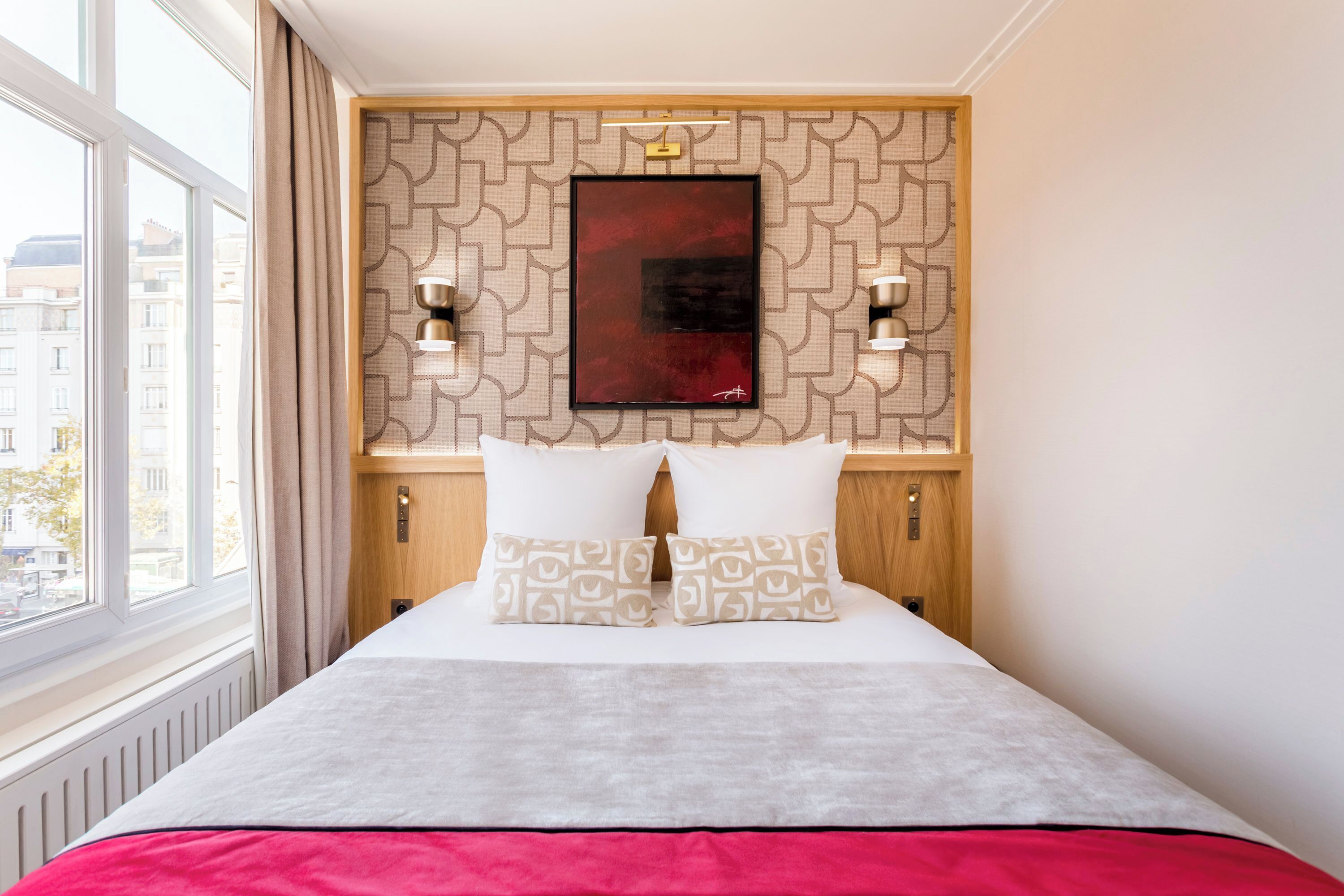 A hotel room with a large bed, a window with curtains, a patterned wall, and two wall-mounted lights.