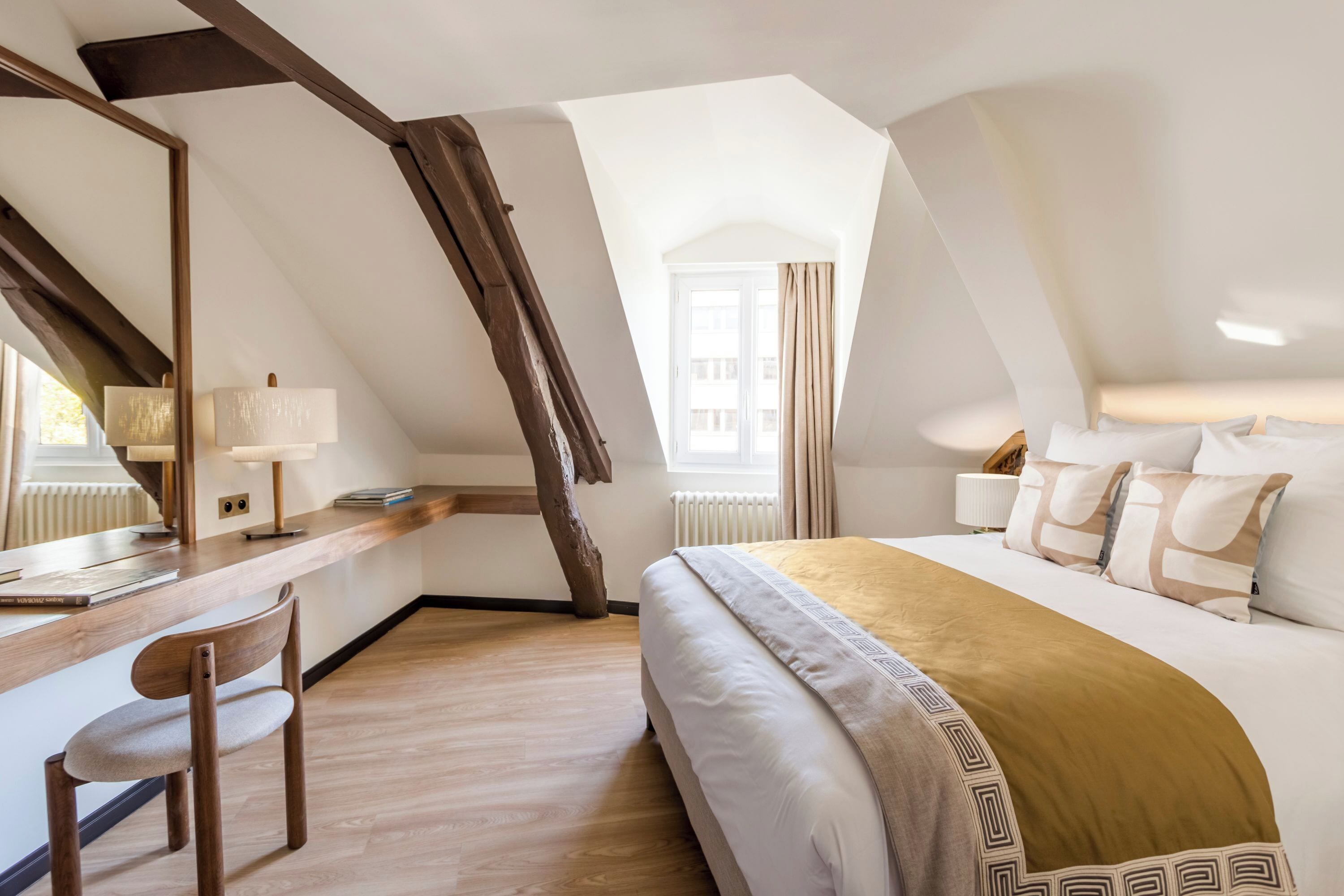 A bedroom with a sloped ceiling, a bed with a patterned throw, a desk with a chair, and a window with curtains.