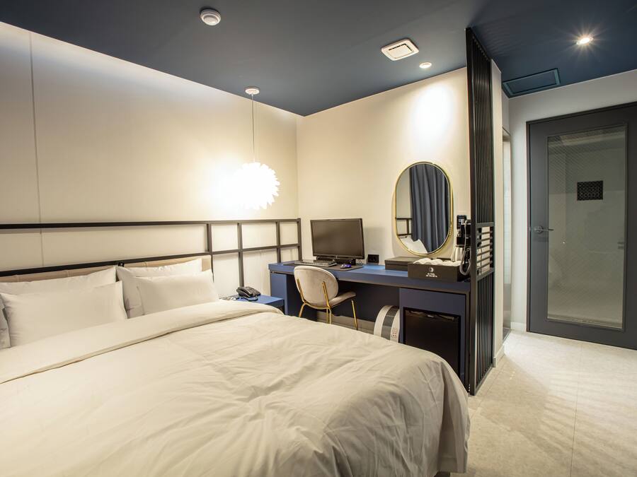 A modern hotel room with a large bed, a desk with a computer, a chair, a mirror, and a door.