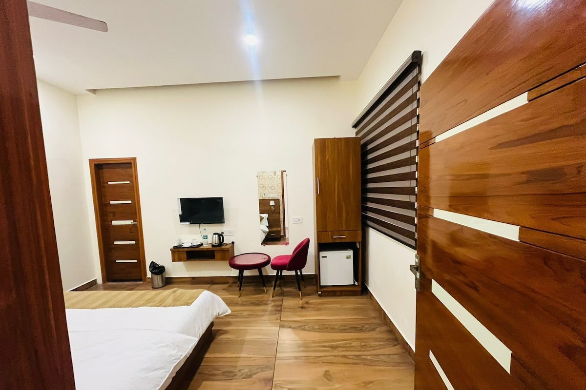 A hotel room with a bed, a desk with a chair, a television, a small refrigerator, and a wardrobe.