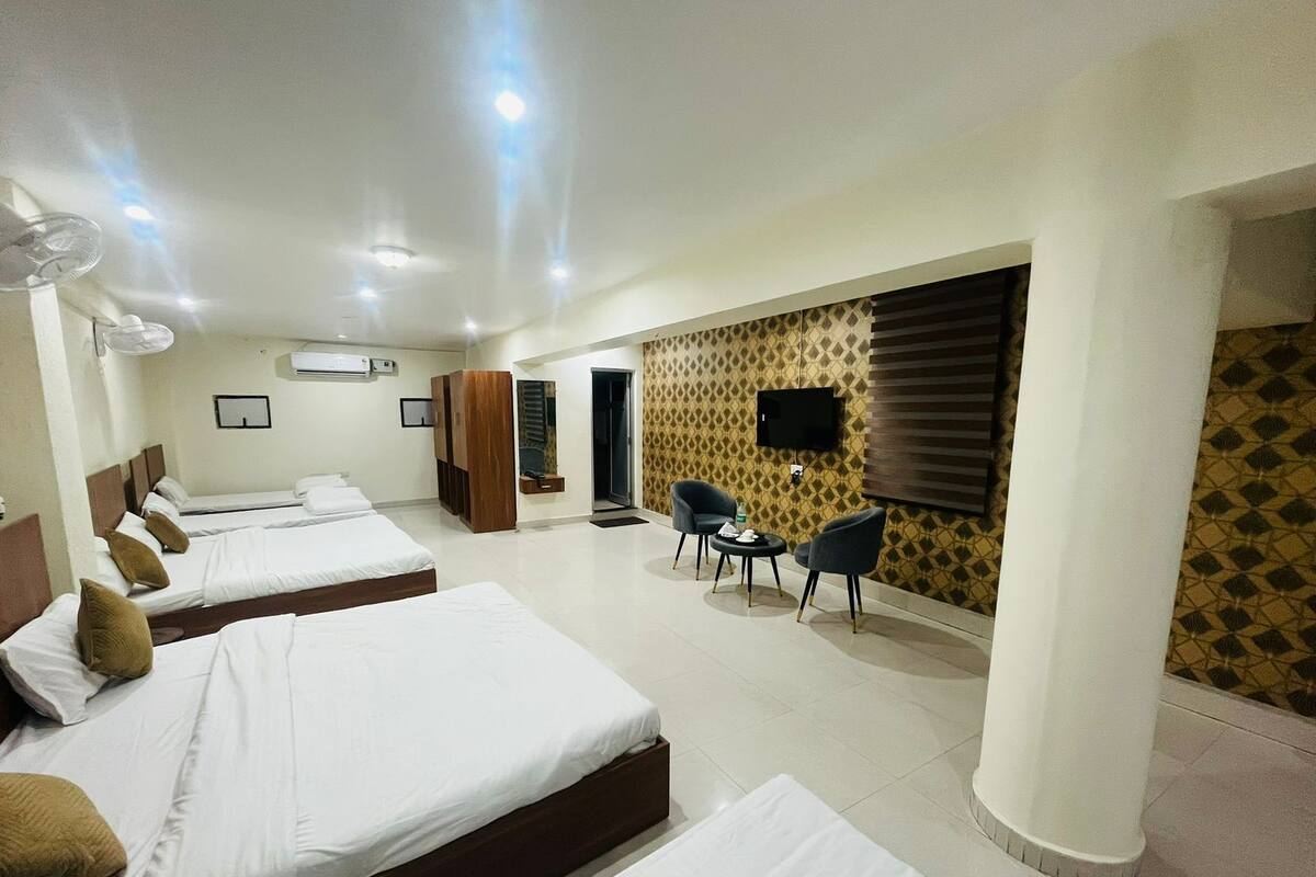 A hotel room with four beds, a TV, a seating area with two chairs, and a patterned wall.