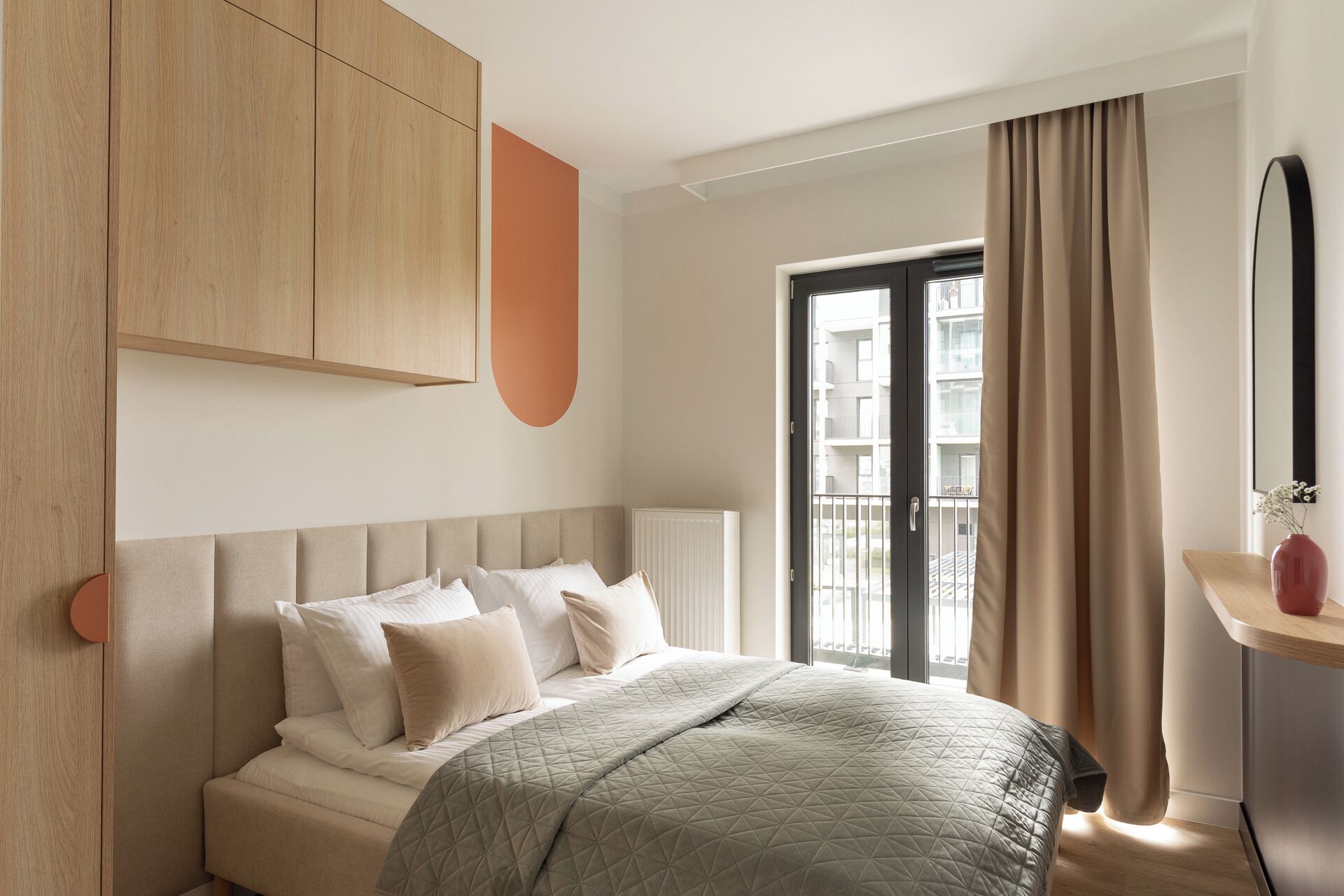 A modern bedroom with a large bed, a wooden wardrobe, a window with curtains, and a small table with a vase.