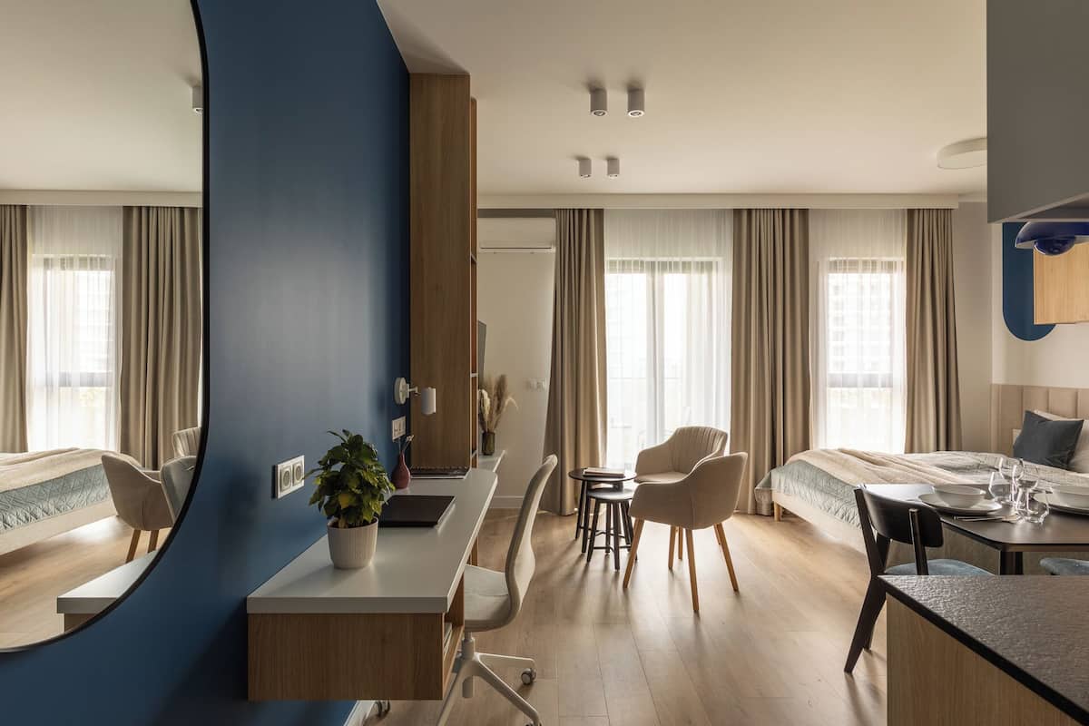 A modern hotel room with a central dining area, a bed with a view, and a large window with curtains.