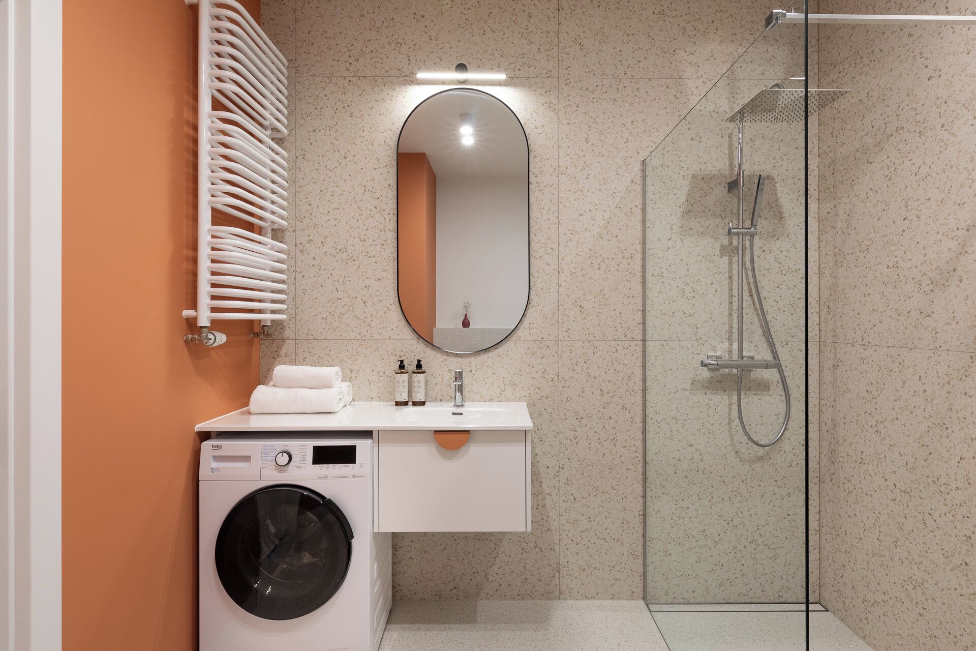 A modern bathroom with a washing machine, a sink with toiletries, a mirror, and a glass-enclosed shower.