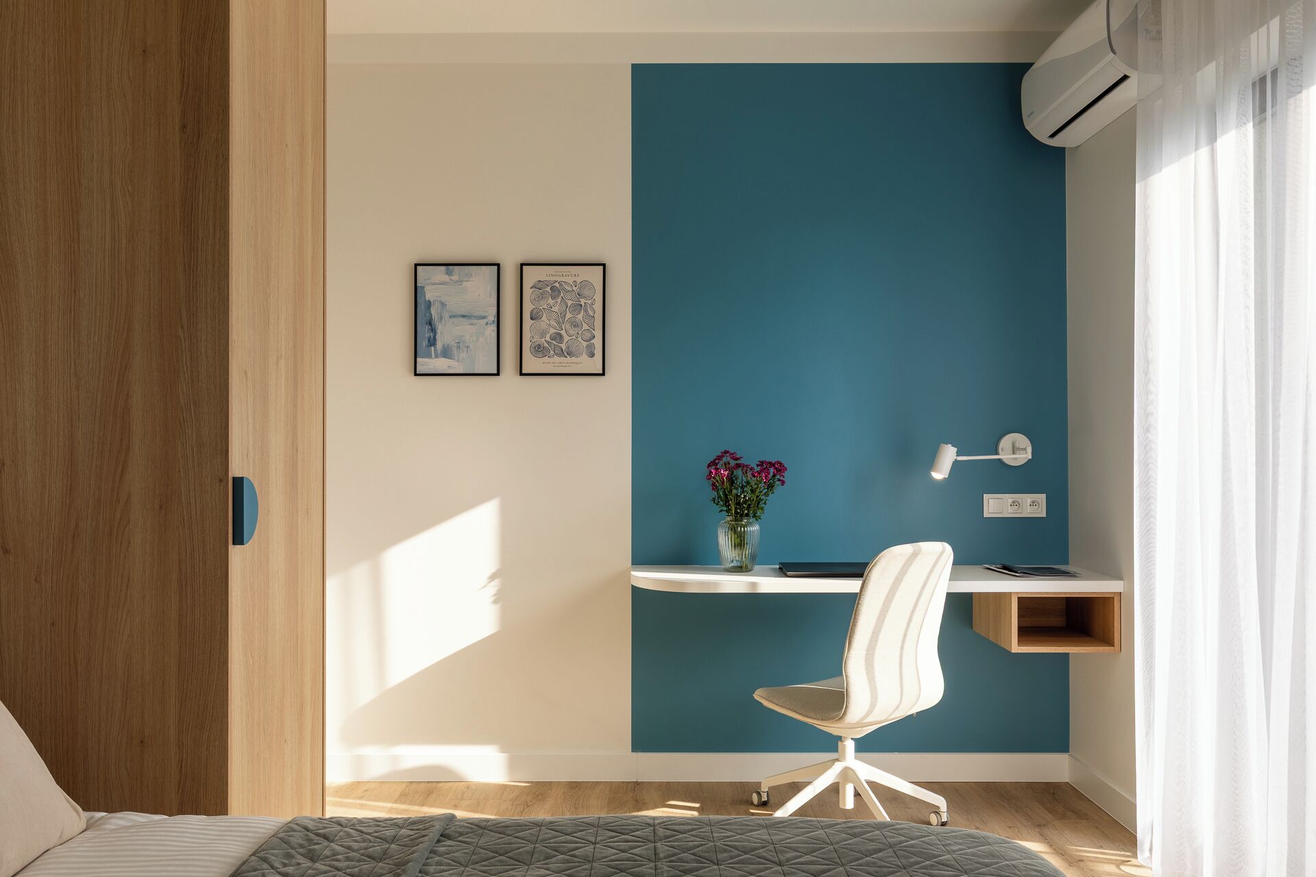 Ein modernes Schlafzimmer mit einer Türkis-farbenen Akzentwand, einem Schreibtisch mit Stuhl, einer Vase mit Blumen und zwei gerahmten Kunstwerken.