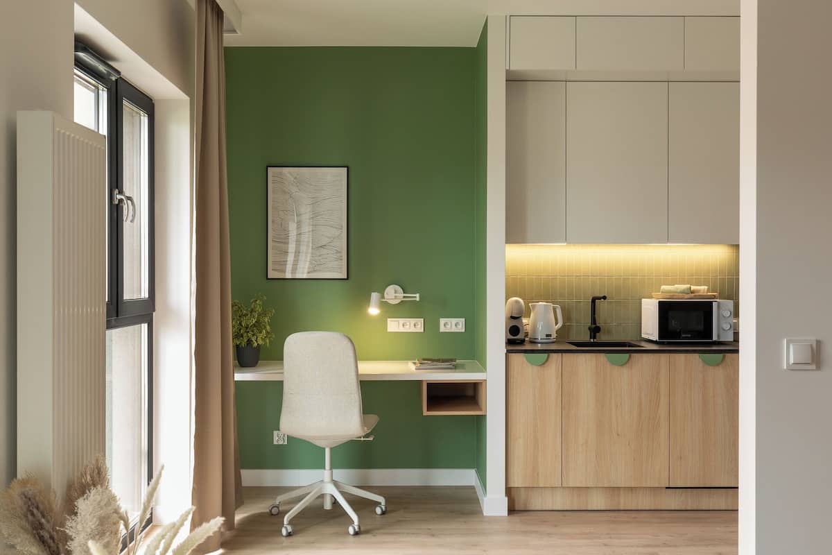 A compact kitchen with a microwave, kettle, and a small sink. A desk with a chair is positioned against a green wall, and a radiator is visible on the left.