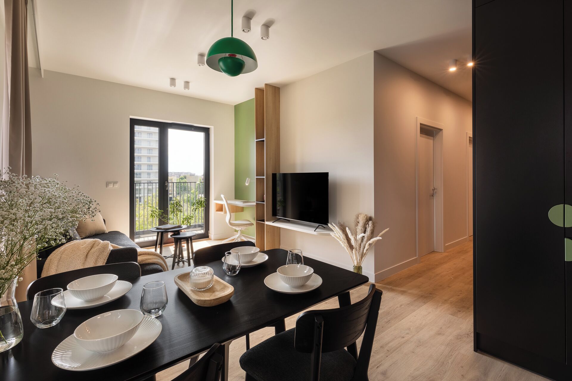 Un espace repas moderne avec une table noire dressée pour quatre personnes, un téléviseur fixé au mur et un balcon offrant une vue sur la ville.