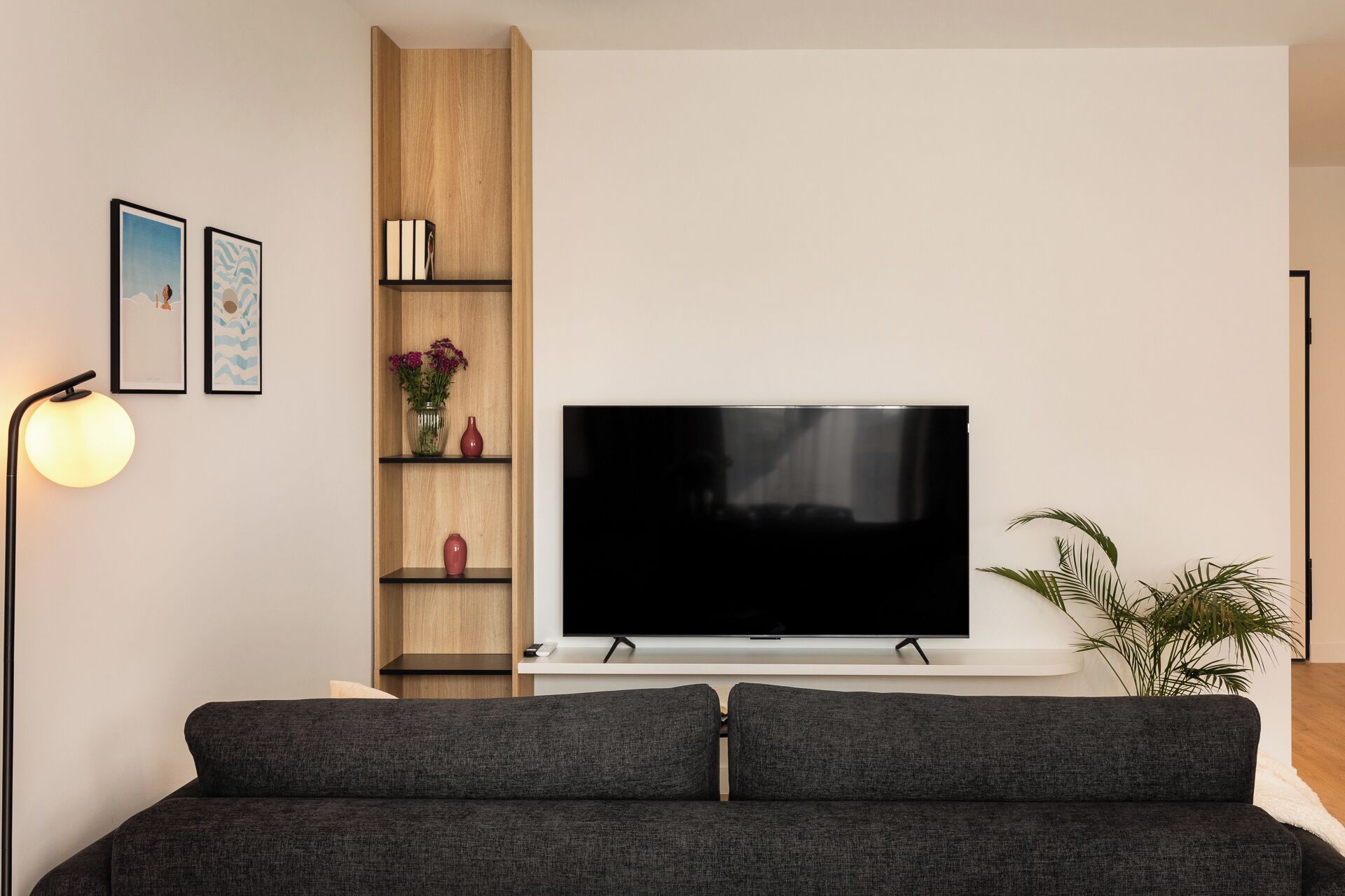 Una sala de estar moderna con un televisor de pantalla plana, un sofá gris oscuro, estanterías de madera con adornos y una lámpara de pie.