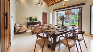 Un comedor con mesa de madera dispuesta para seis personas, rodeada de sillas. Un televisor está montado en la pared, y una ventana ofrece vistas a un jardín.