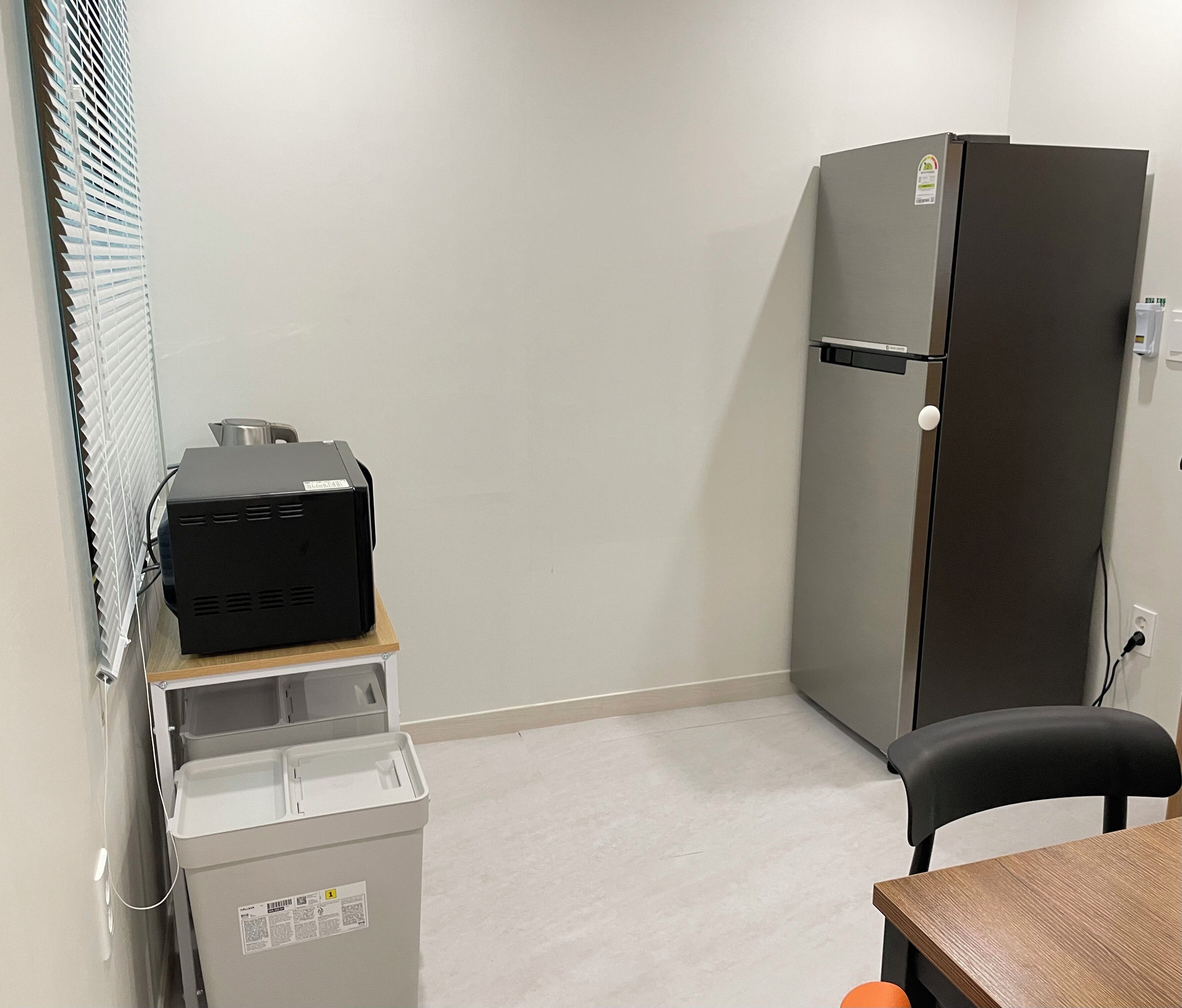 A compact room with a refrigerator, a small table, a chair, and a trash bin.