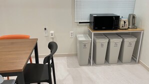 Ett litet pentry med en mikrovågsugn, brödrost och kaffebryggare på en hylla av trä. Det finns sopkärl märkta för olika typer av avfall: hushållssopor, burkar, glas och plast. En svart stol och ett träbord syns.