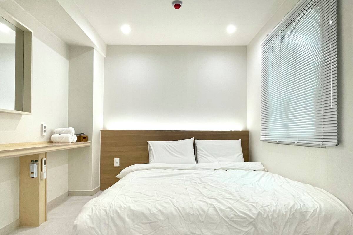 Ein modernes Hotelzimmer mit einem großen Bett, einem Holz-Kopfteil, einem Nachttisch mit Spiegel und einem Fenster mit Jalousien.