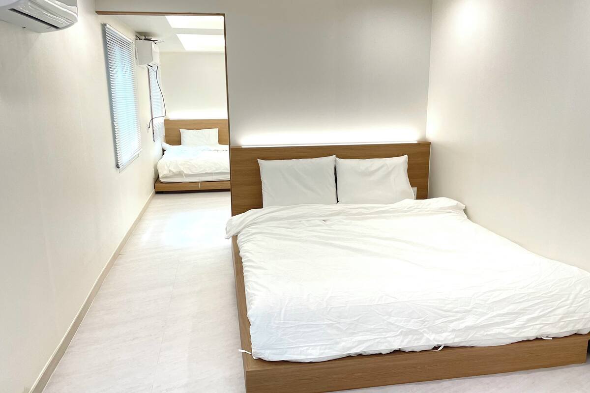 Ein modernes Hotelzimmer mit einem Bettgestell aus Holz, weißer Bettwäsche und einem großen Fenster mit Jalousien.