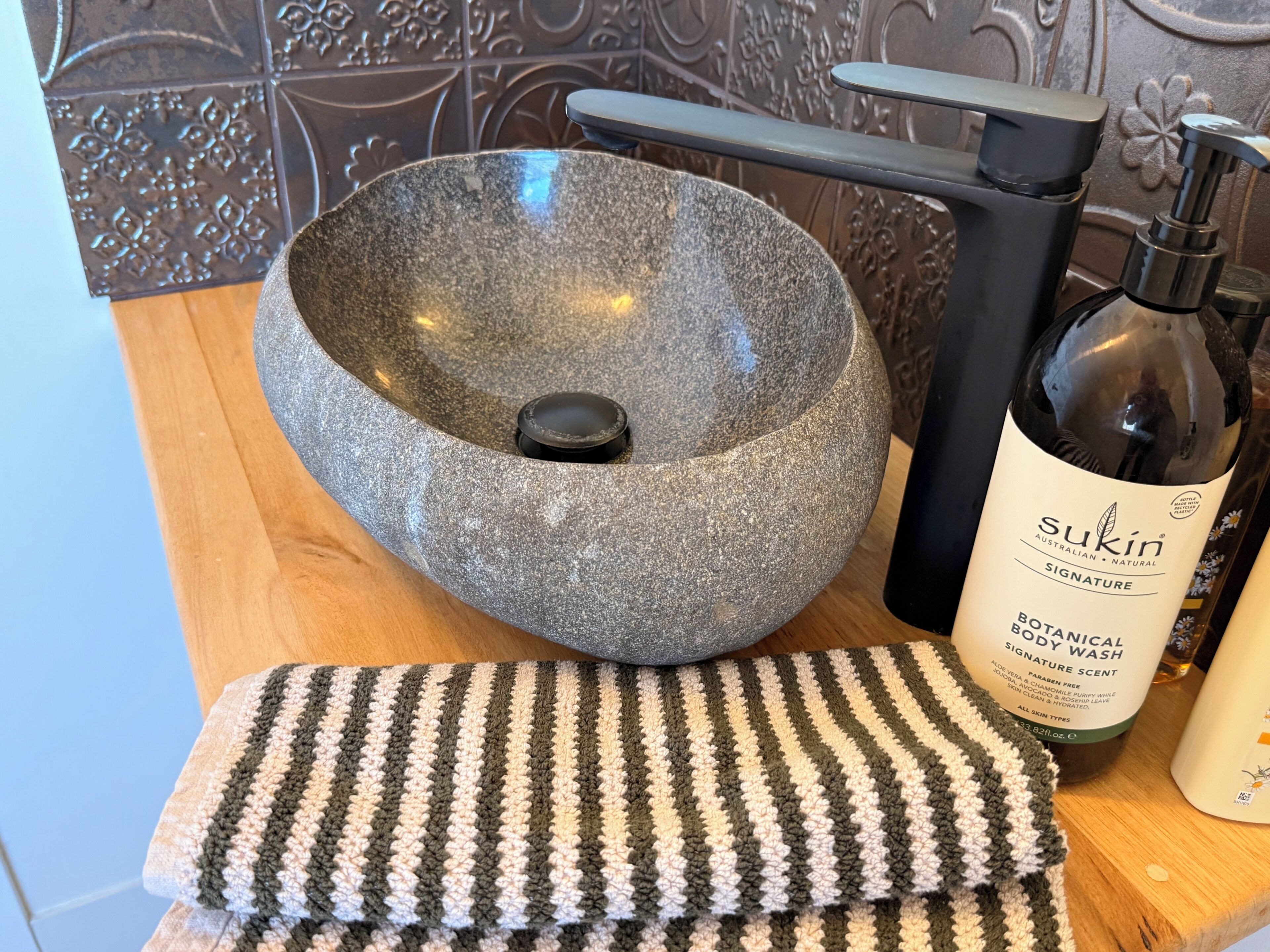 A stone basin, a bottle of Sukin Signature Botanical Body Wash, and a striped towel on a wooden surface.