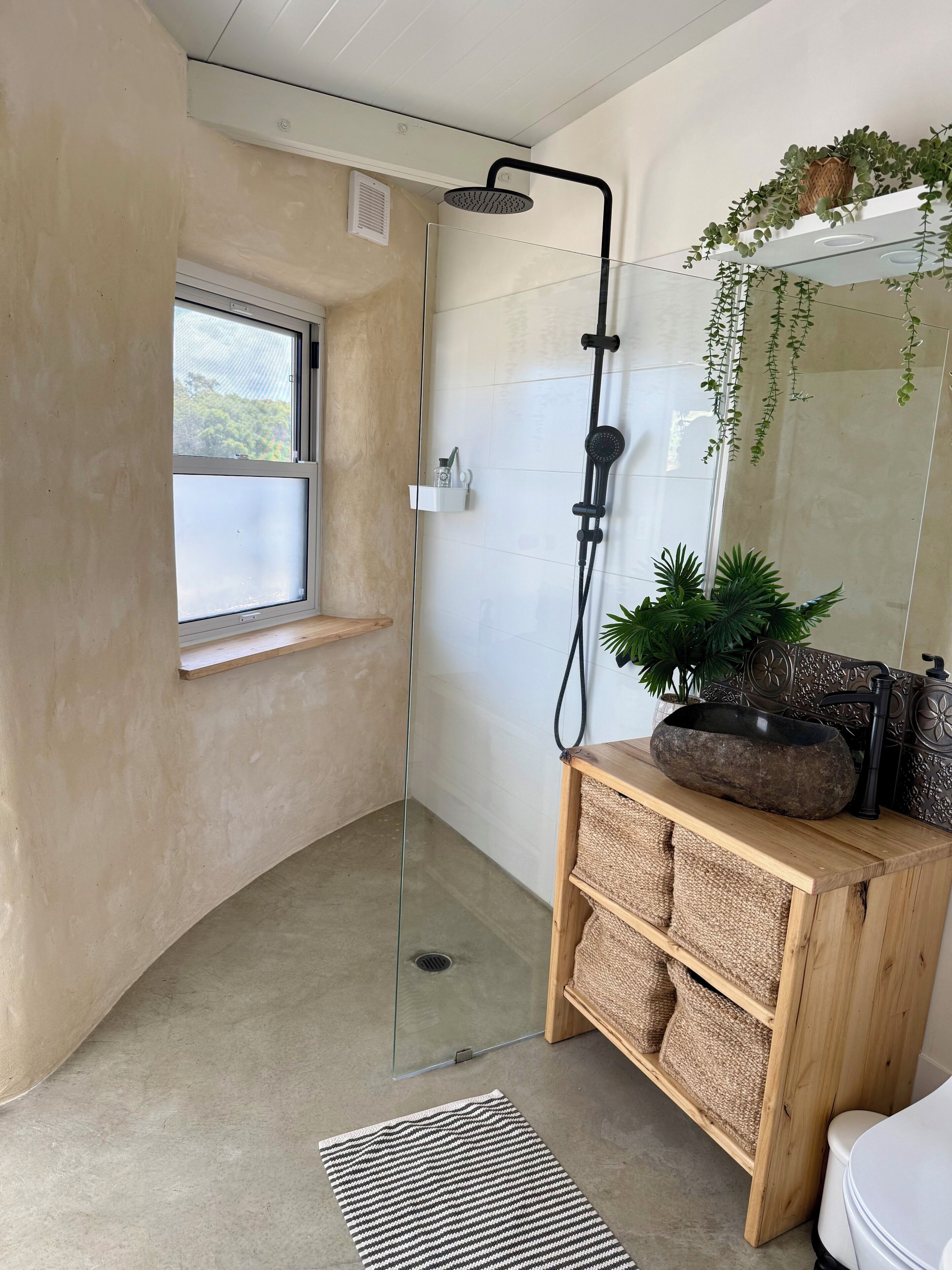 A modern bathroom with a glass shower enclosure, a wooden vanity with a black sink, and a window with a view of greenery.