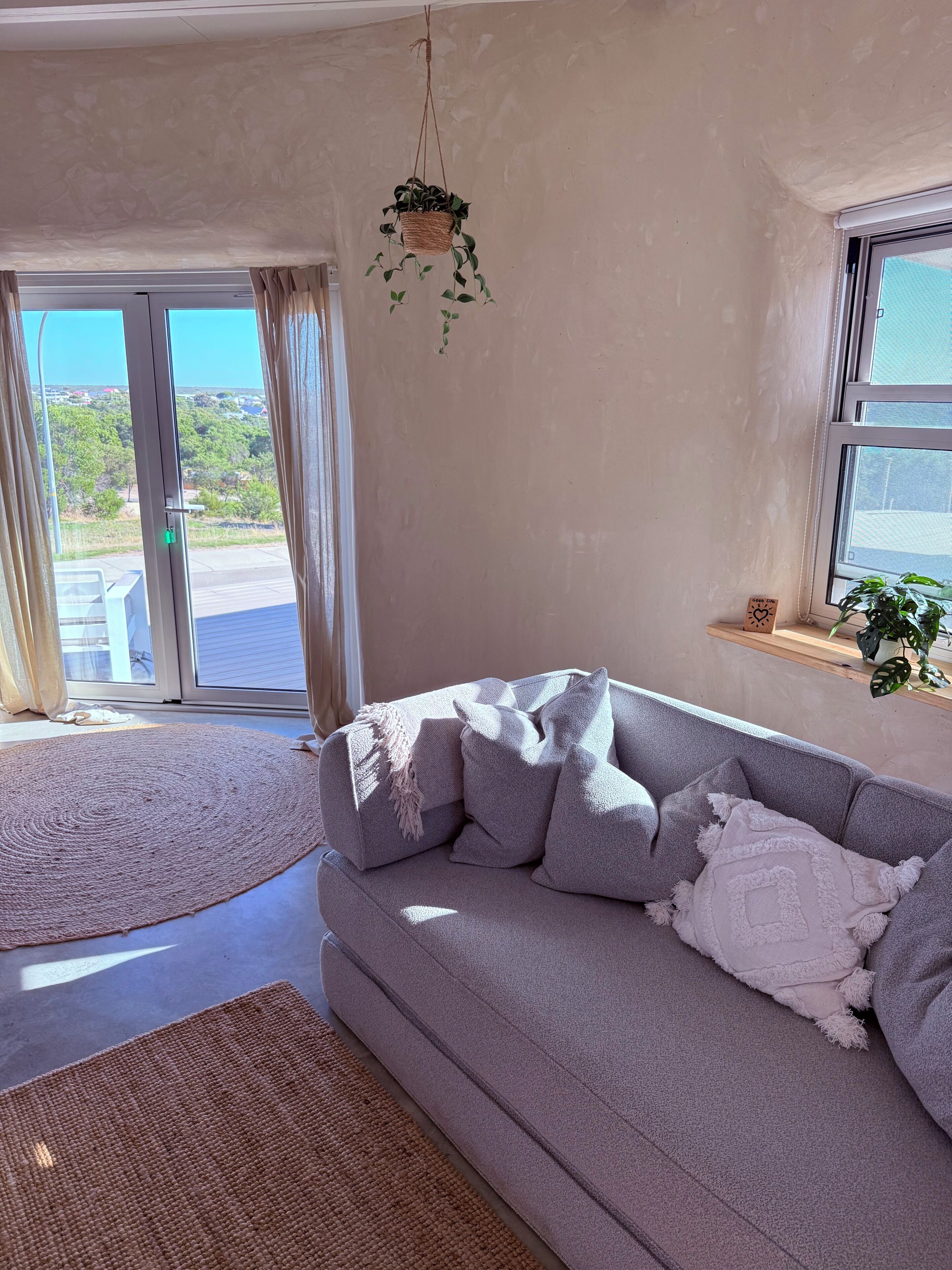 A living room with a grey sofa, a woven rug, a hanging plant, and a view of the outdoors through a glass door.