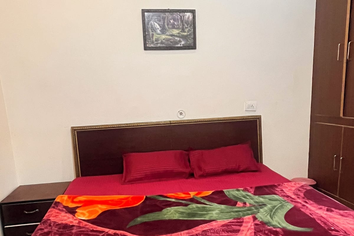 A bed with a red and orange floral bedspread, a dark brown headboard, and a small framed picture on the wall above.
