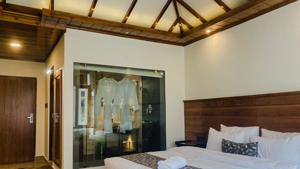 A bedroom with a large bed, a wooden headboard, a bench, and a glass-enclosed walk-in closet with robes. - Namdul Retreat (Mangan)