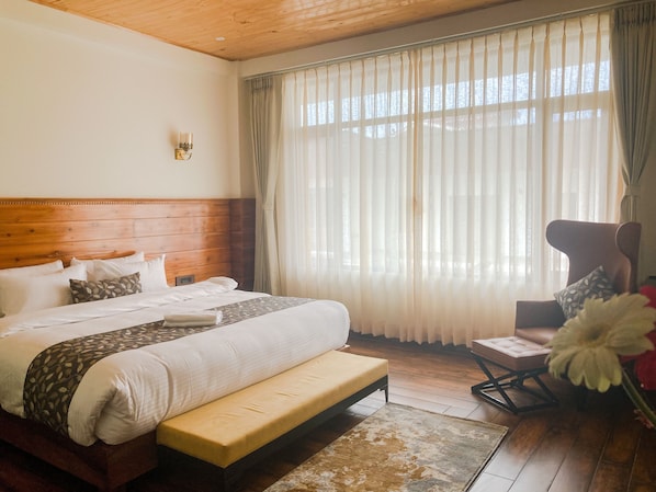 A hotel room with a wooden headboard, a large bed with white linens, a bench, a chair, and a large window with sheer curtains. - Namdul Retreat (Mangan)