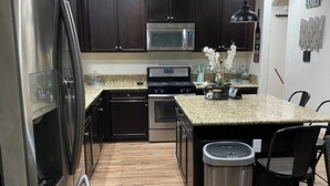 A modern kitchen with dark wooden cabinets, stainless steel appliances, a granite countertop, and a dining area with a black chair. - 4-bedroom house in an amazing neighborhood with AC, WiFi. Relax and unwind. (Imperial)