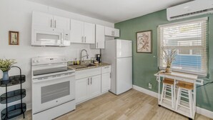 A kitchen with white cabinets, a stove, a microwave, and a refrigerator. There is a window with blinds and a small table with a plant. - 2528 Renovated Studio Near The Beach (Melbourne)
