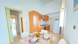 A compact kitchen with wooden cabinets, a white refrigerator, and a dining area with a floral tablecloth. - Beautiful home with panoramic sea view (borders state property) (Marina di Mancaversa)