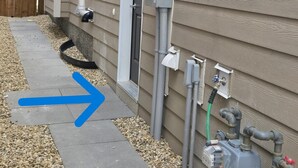 Fachada de una casa con cajas eléctricas exteriores, un calentador de agua y una flecha direccional en el suelo.