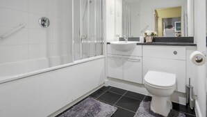 Egy modern fürőszoba, üvegfalas zuhanyzóval, fehér WC-vel és egy mosdókagylóval, mely felett tükör található.