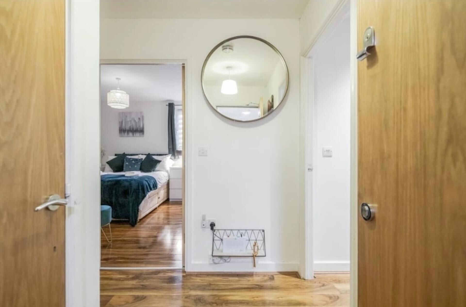 Un couloir mène à une chambre dotée d’un plancher en bois, d’un miroir rond accroché au mur et d’un lit avec une tête de lit bleue.