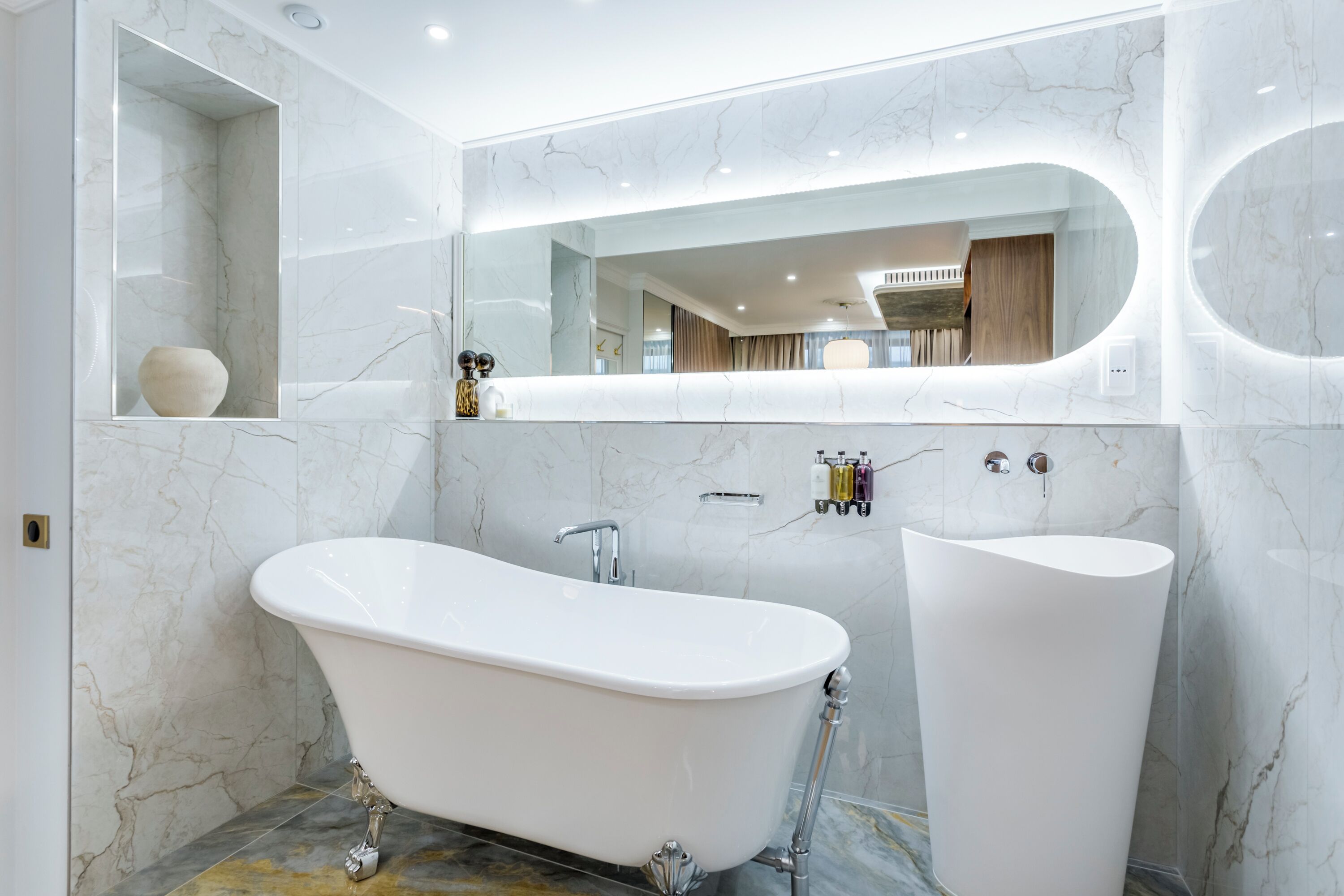 A modern bathroom with a freestanding bathtub, a large mirror, and marble walls.