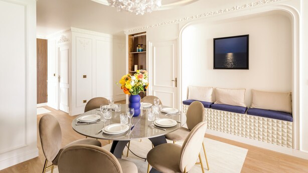 Un comedor con una mesa redonda dispuesta para seis personas, rodeada por seis sillas. Hay un jarrón con flores sobre la mesa, y una estantería con libros y objetos decorativos al fondo.