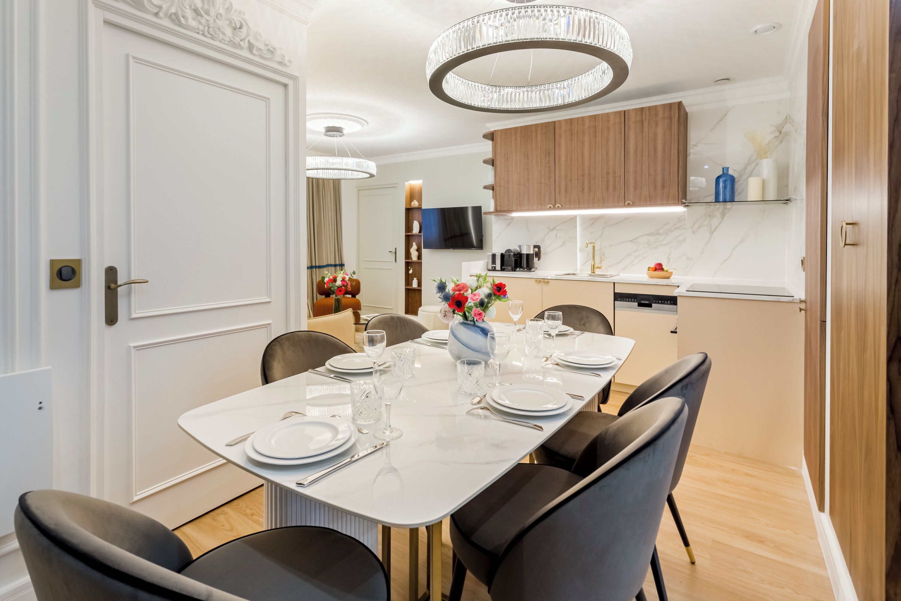 A modern dining area with a white table set for six, surrounded by black chairs. A chandelier hangs above, and wooden cabinets are visible in the background.
