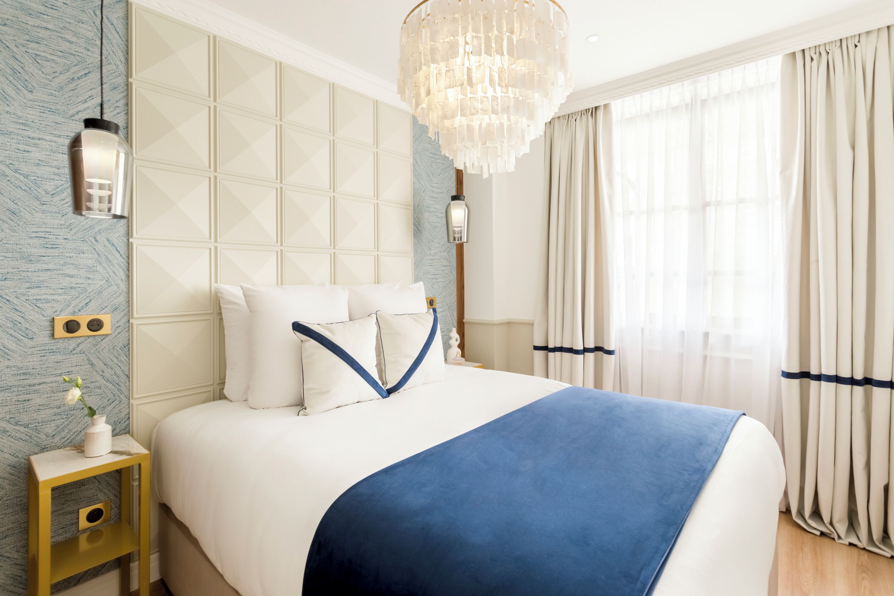 A modern bedroom with a large bed, a chandelier, and a view of the outdoors through sheer curtains.