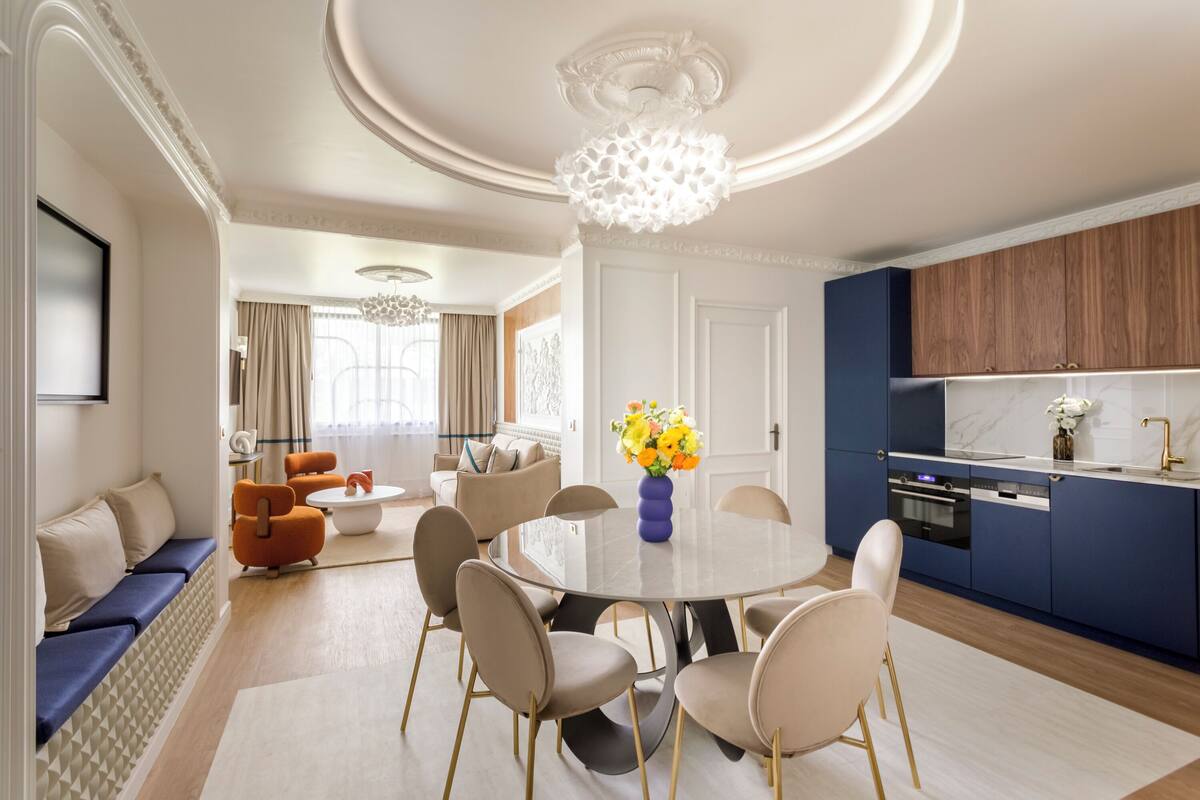 Un salon moderne avec une table de salle à manger ronde, des canapés de couleur beige et un coin cuisine doté de meubles bleus.