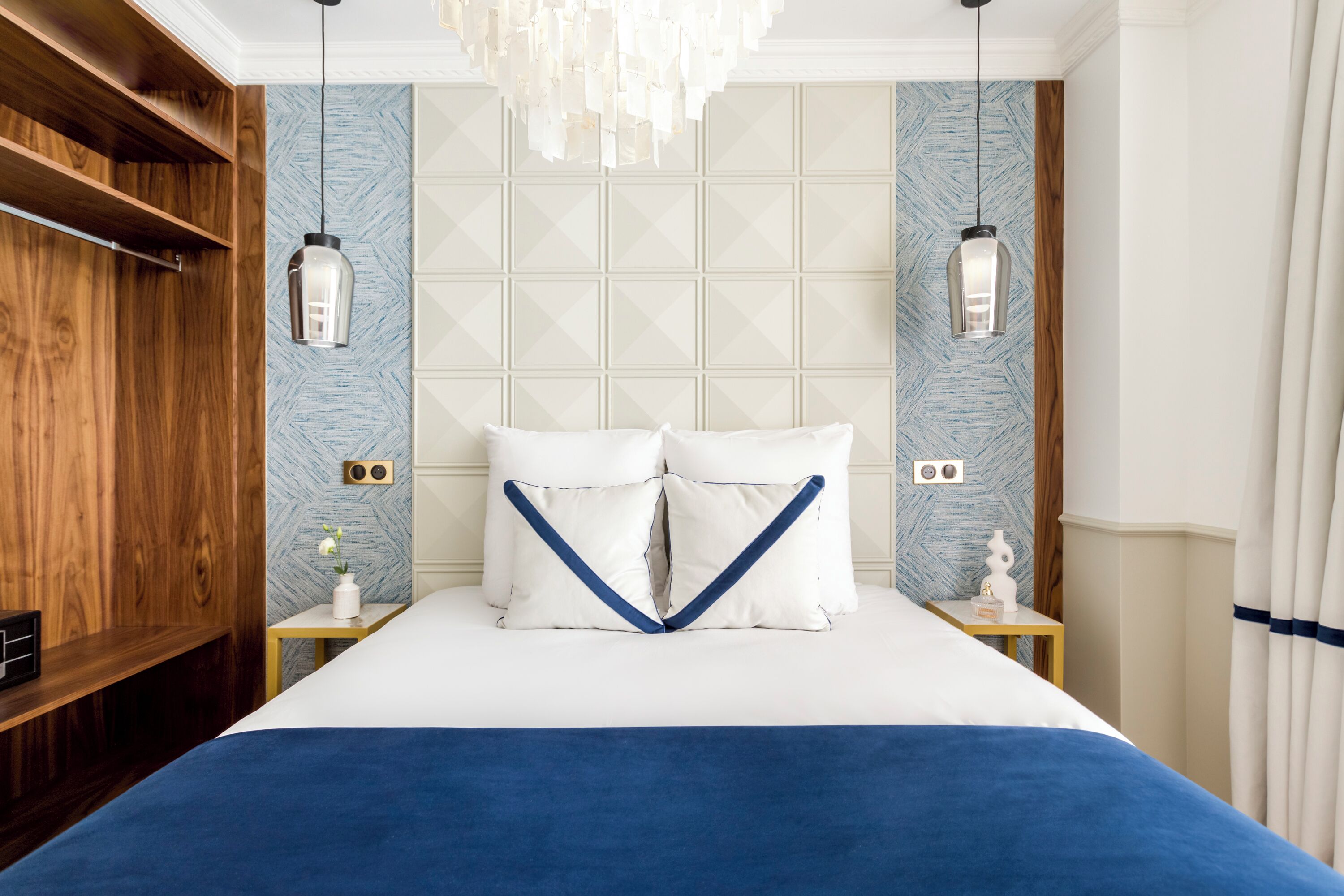 A neatly arranged hotel room with a large bed, wooden paneling on one side, and a geometric patterned wall.